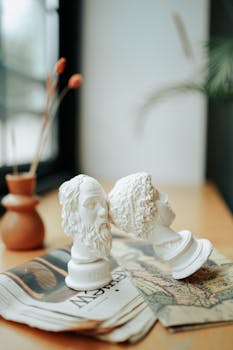 Two classical bust sculptures on a table with newspapers and artistic decor.